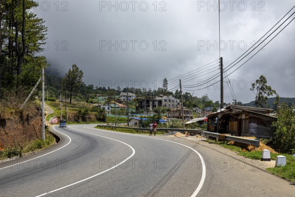 A winding road in a rural area with buildings, trees and a cloudy sky, road through the mountains near Kandy in Sri Lanka