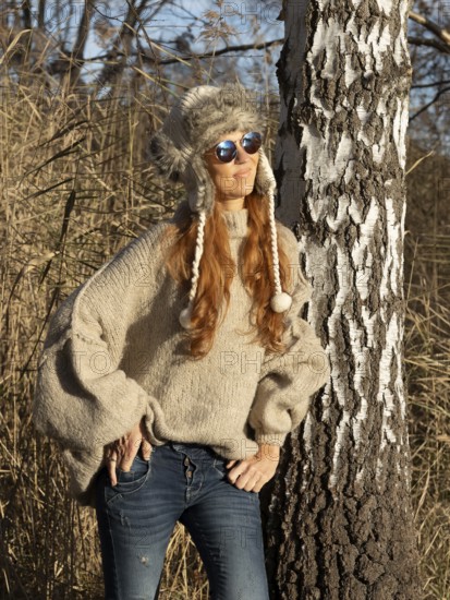 Slender pretty woman with fur hat and pompoms, red hair and warm winter colors between birch forest and reeds