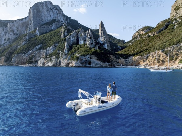 Couple on motorboat off picturesque rocky coast, cliffs with L'Aguglia pinnacle, blue sea and Cala Goloritzé beach, aerial view, Golfo di Orosei, Baunei, Sardinia, Italy