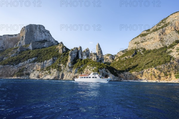 Excursion boat off picturesque rocky coast, cliffs with caves in morning light, L'Aguglia rock pin, blue sea and Cala Goloritzé beach, Golfo di Orosei, Baunei, Sardinia, Italy
