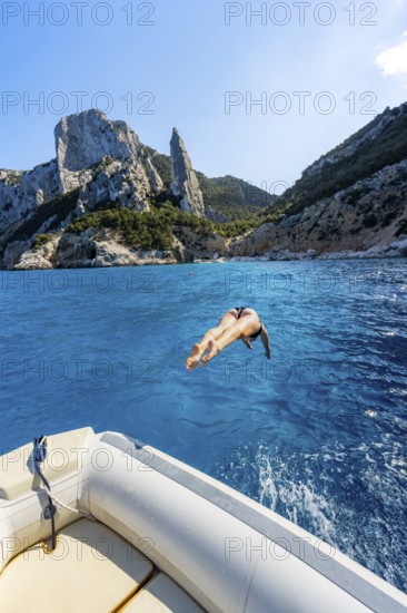 Young woman jumping from a boat into the water, picturesque rocky coast, cliffs with L'Aguglia rock pin, blue sea and Cala Goloritzé beach, Golfo di Orosei, Baunei, Sardinia, Italy