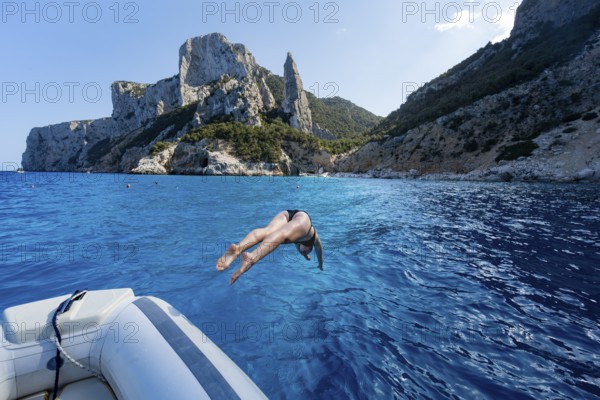Young woman jumping from a boat into the water, picturesque rocky coast, cliffs with L'Aguglia rock pin, blue sea and Cala Goloritzé beach, Golfo di Orosei, Baunei, Sardinia, Italy