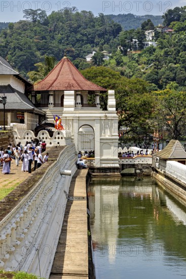 A temple with a pavilion near a river, with people and green surroundings, The Temple of the Tooth in Kandy Sri Lanka