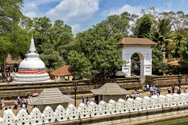 A temple complex with a white stupa surrounded by trees and people under clear skies, The Temple of the Tooth in Kandy Sri Lanka