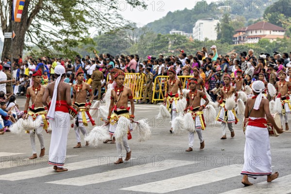 Children move across a street in traditional costumes, spectators watch, The Perahera Parade in Kandy Sri Lanka