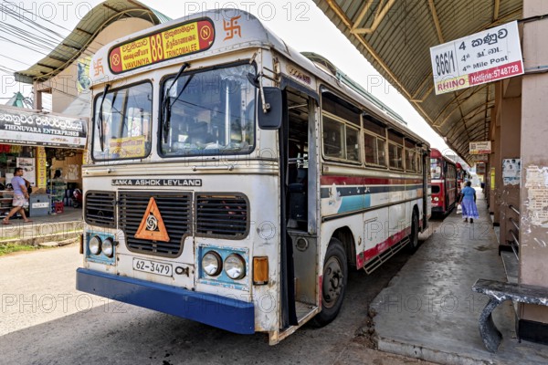 An Ashok Leyland bus at a busy bus stop in Sri Lanka, surrounded by signs and passers-by, bus station in Kandy Sri Lanka