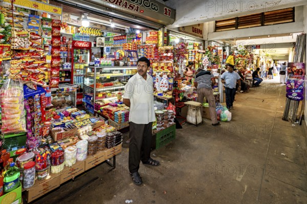 A man stands in a well-stocked shop with numerous colorful foods and drinks, dealers in the market halls of Kandy Sri Lanka