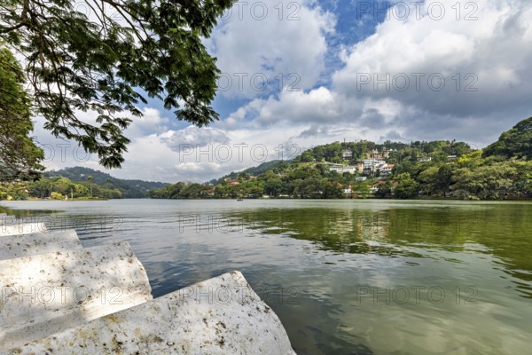 Painting of a calm lake surrounded by green hills and trees under a partly cloudy sky, Lake Kandy in Kandy Sri Lanka