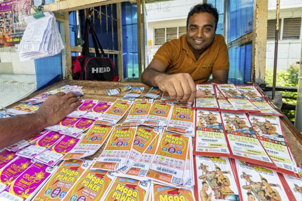 A man smiling behind a sales desk with colorful lottery tickets and tickets, dealer in the market halls of Kandy Sri Lanka