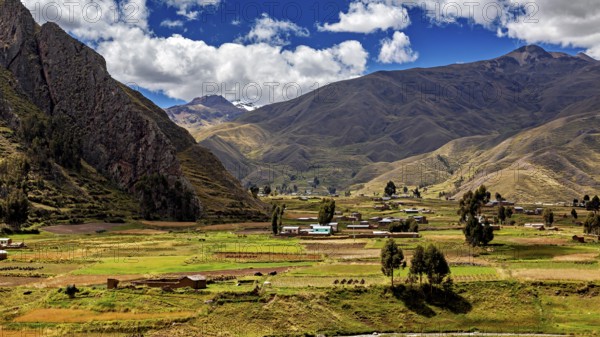 Green fields and hills against a mountain backdrop under a cloudy blue sky, the landscape in the highlands of Peru