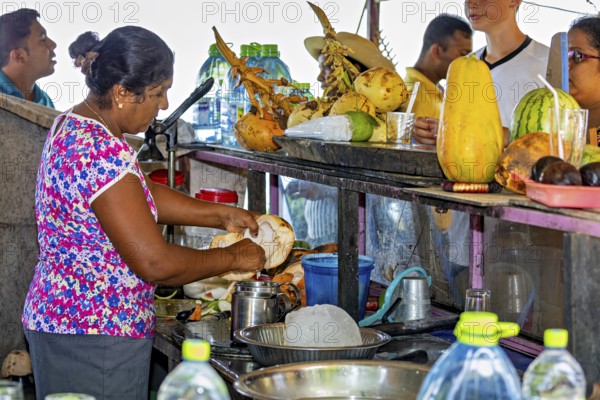 Woman prepares coconuts at a busy market stall surrounded by people and tropical produce, traditional small beach bar on Trincomalee beach in Sri Lanka
