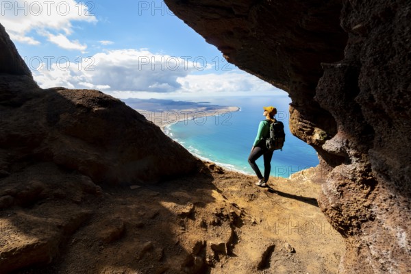 Tourist in the Cueva de las Cabras cave, young woman enjoying the view from the Risco de Famara cliff to Famara beach, Playa de Famara with La Calaeta, Lanzarote, Canary Islands, Spain