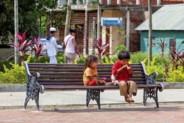 Two children sit on an outdoor bench while they eat. The surrounding area is green and peaceful, two children on a park bench in Rurrenabaque in the Amazon jungle rainforest in Bolivia
