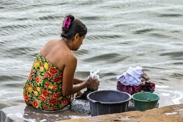 A woman is sitting by the river washing clothes with several buckets, a woman is washing at a lake near Sigiriya in Sri Lanka