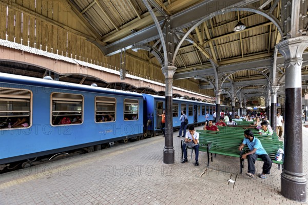 People sitting on green benches in the train station, blue train on the platform, covered structure, the station and platform with trains from Colombo in Sri Lanka