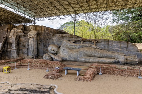 Panoramic view of a large reclining Buddha statue with a figure standing next to it under a protective canopy, The Buddhas near Polonnaruwa in Sri Lanka