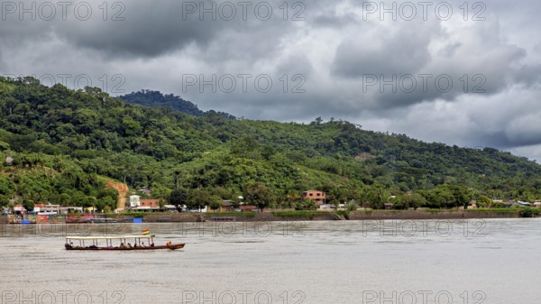 A boat moves on a river in front of a wooded hill and under a cloudy sky, people on boats near Rurrenabaque in the Amazon jungle in Bolivia