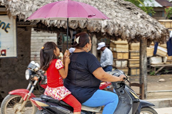 A woman and a child ride on a moped under a red umbrella in a tropical setting, A woman and child riding on a moped in the streets of Rurrenabaque in Bolivia