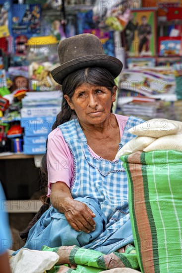 A thoughtful woman in traditional traditional costume sits in front of a colorful market stall, The Rurrenabaque Market in Bolivia