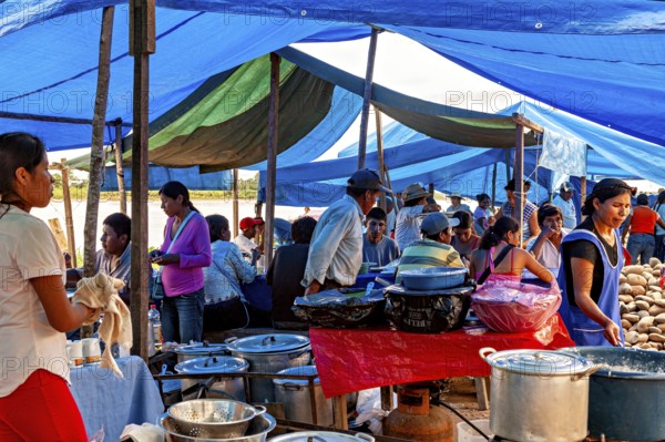 People cook and serve food under blue tarps in a busy market atmosphere, The Rurrenabaque market in Bolivia