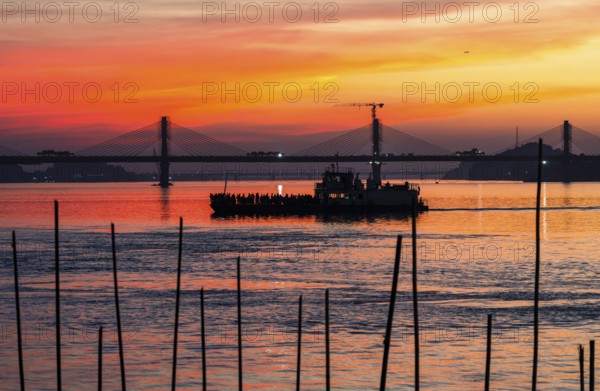 A boat sails on the river Brahmaputra during sunset, with the silhouette of a cable-stayed bridge and hills in the background, on November 4, 2025 in Guwahati, India