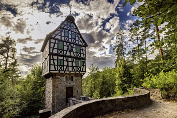 A tall, narrowly built half-timbered house on a stone base surrounded by dramatic skies with lush vegetation, The Herzogstuhl in the Saale Holzlandkreis district in Thuringia