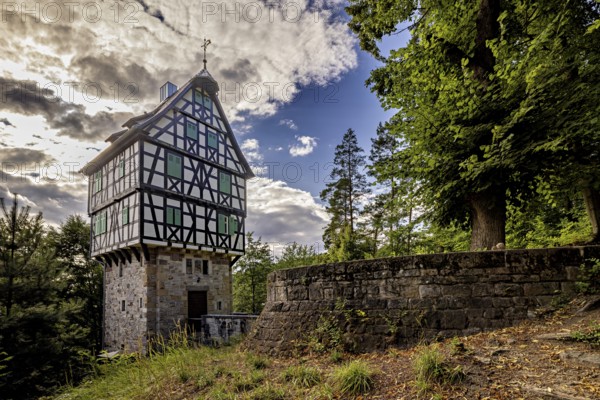 Half-timbered house on a stone base next to a stone wall in the forest, picturesquely illuminated by the sun with dramatic clouds, The Herzogstuhl im Saale Holzlandkreis in Thuringia