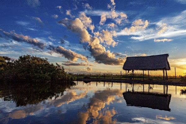 A thatched hut reflected in the water at sunset with dramatic cloud formations, The pampas swamps in the Amazon basin of Bolivia