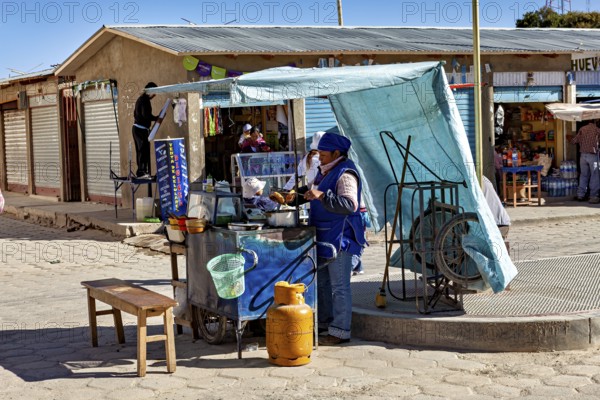 Street snack bar under a blue sunshade, woman serving customers next to a table and gas canister in a sunny environment, Small shops in the town of Uyuni in the Salar de Uyuni in Bolivia