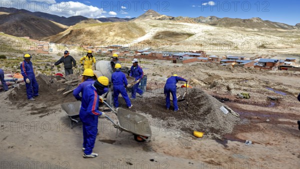 Workers in blue work clothes shovel earth on a sunny hilly landscape, surrounded by mountains and houses, workers on the roadside on the Altiplano in Bolivia