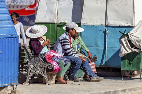 People with hats sitting on a bench in the sun at a market stall, people of the city of Uyuni in the Salar de Uyuni in Bolivia
