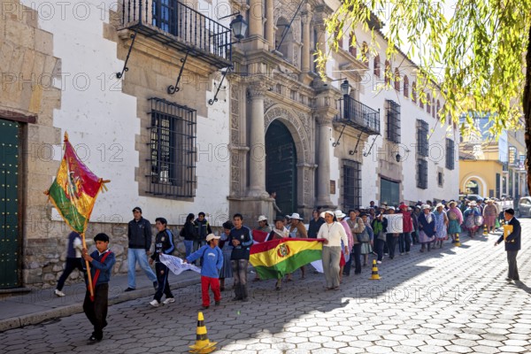 People march in a parade through a sunny street with historic buildings and flags, demonstration in the streets of Potosi Bolivia