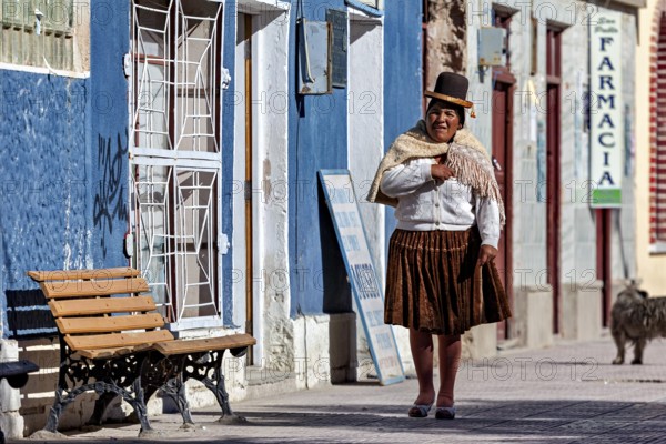Woman with a traditional hat stands in front of a blue house wall in the sun, people of the city of Uyuni in the Salar de Uyuni in Bolivia