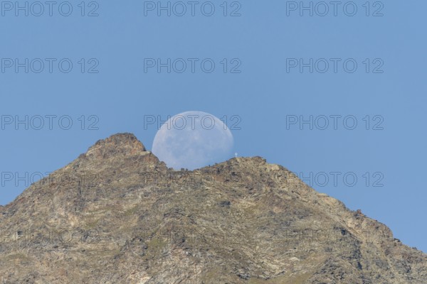 Majestic mountain peak stands tall against vibrant blue sky as large moon rises just behind it. The scene captures the quiet beauty of nature in the early evening light. Grachen, Viege, Valais, Switzerland