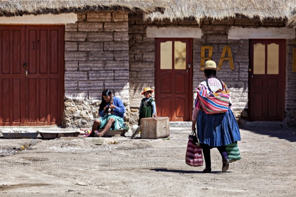 A woman in traditional dress carries bags while children and another person sit in the background, Traditional woman in a village in the Salar de Uyuni in Bolivia