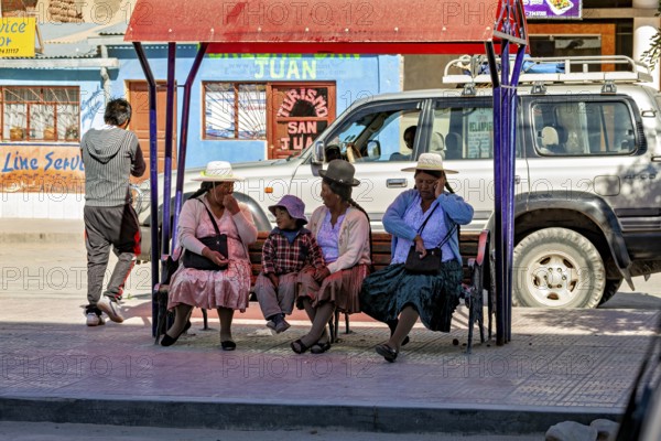 People in traditional dress sit on a bench in an urban environment as a car drives past, people at a bus stop in Uyuni, Bolivia