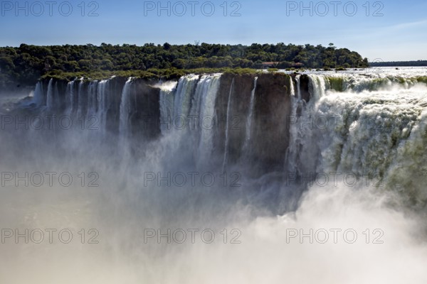 Thick mist rises from the mighty waterfall, flanked by green forest under a blue sky, The Iguazu Falls between Argentina and Brazil