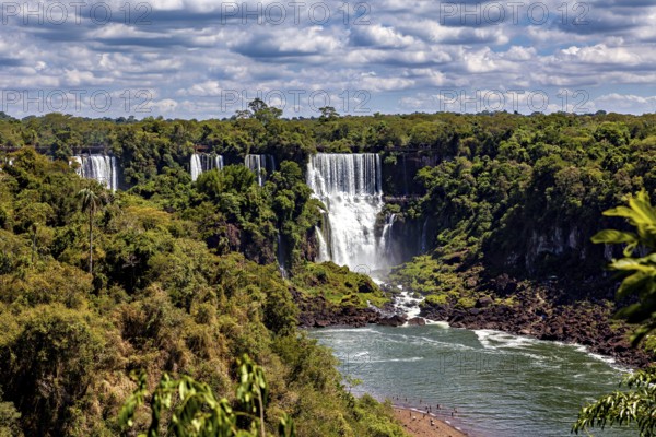 Large waterfalls flow into a river, surrounded by lush vegetation under a cloudy sky, The Iguazu Falls between Argentina and Brazil
