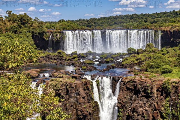 Steep waterfalls cascade over rocks, surrounded by dense greenery and a cloudy sky, The Iguazu Falls between Argentina and Brazil