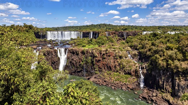 Large waterfalls flow over steep cliffs, surrounded by lush greenery and a blue sky with clouds, The Iguazu Falls between Argentina and Brazil