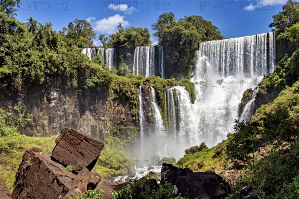 Waterfalls cascade through lush vegetation, surrounded by rocks under a blue sky, The Iguazu Falls between Argentina and Brazil