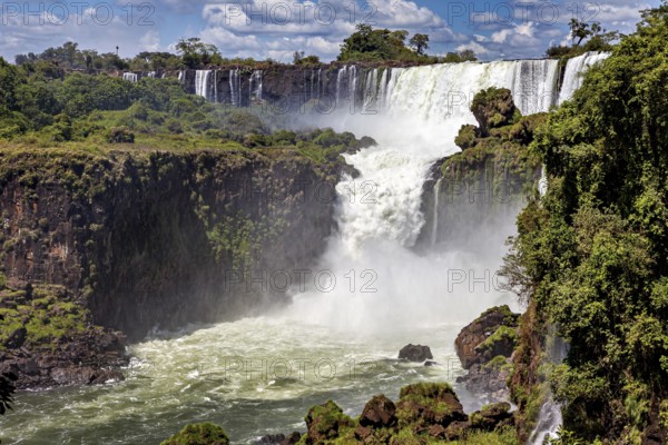 Large waterfall with dense spray and surrounding green forests under a blue sky, The Iguazu Falls between Argentina and Brazil