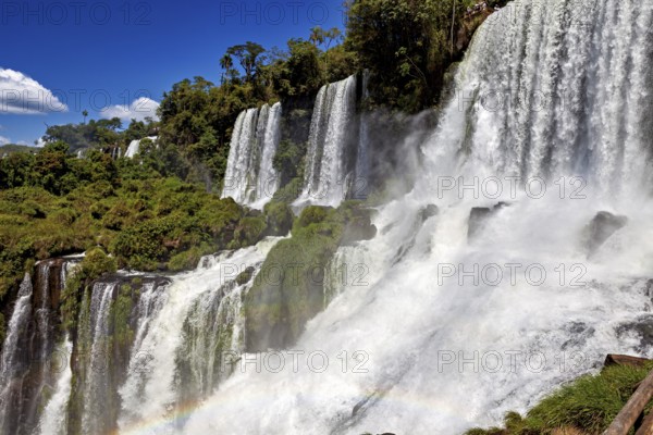 Massive waterfalls create dense spray and a rainbow, surrounded by green forest and blue sky, The Iguazu Falls between Argentina and Brazil