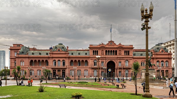 A historic, pink colonial-style building with a cloudy sky and palm trees in the foreground, the Casa Rosada presidential palace in the city of Buenos Aires Argentina