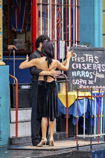 A couple dances tango in front of a colourful building, hugging each other, next to a menu board, The colourful city and tango district La Bocca in Buenos Aires Argentina