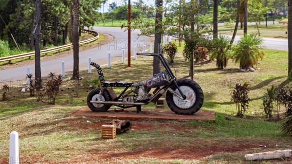 A metal motorbike bust stands in a park with green spaces and trees, placed on a pedestal, A motorbike model on the roadside in Argentina