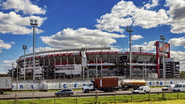 A large stadium with several stands and floodlights under a blue sky and passing vehicles, the River Plate football stadium in Buenos Aires Argentina