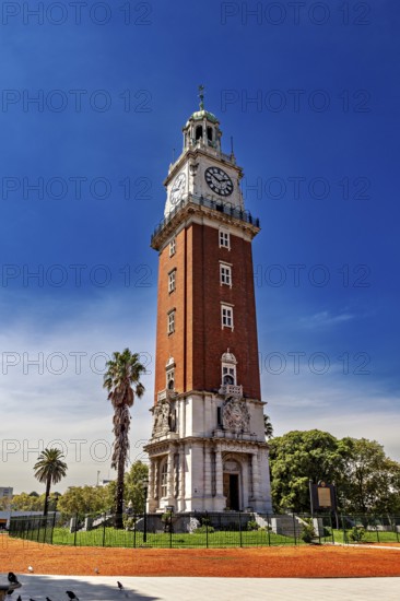 A tall brick clock tower next to palm trees under a clear blue sky, The city of Buenos Aires in Argentina