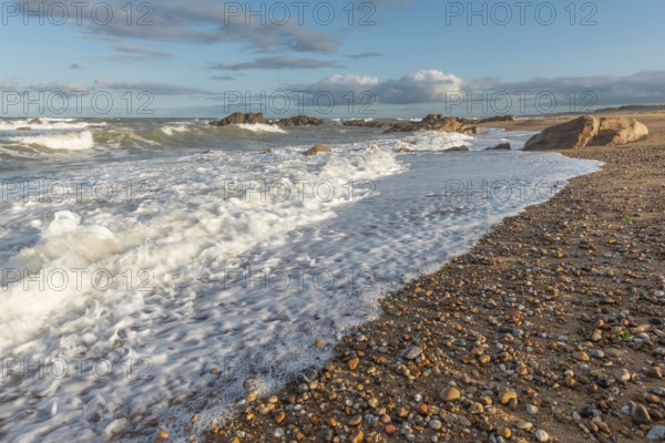 The waves crash gently against a rocky shoreline at sunset, creating a tranquil atmosphere. The sandy beach is strewn with pebbles and surrounded by a beautiful sky. Plage de l'Aubraie, Les Sables d'Olonne, Vendee, France