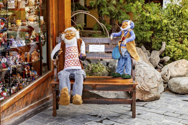 Two dolls sit on a bench in front of a shop window, surrounded by plants and wooden surfaces, dwarves and gnomes from Patagonia in South America